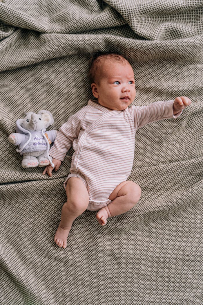 Adorable baby in a striped onesie lies beside a plush elephant toy on a soft blanket.