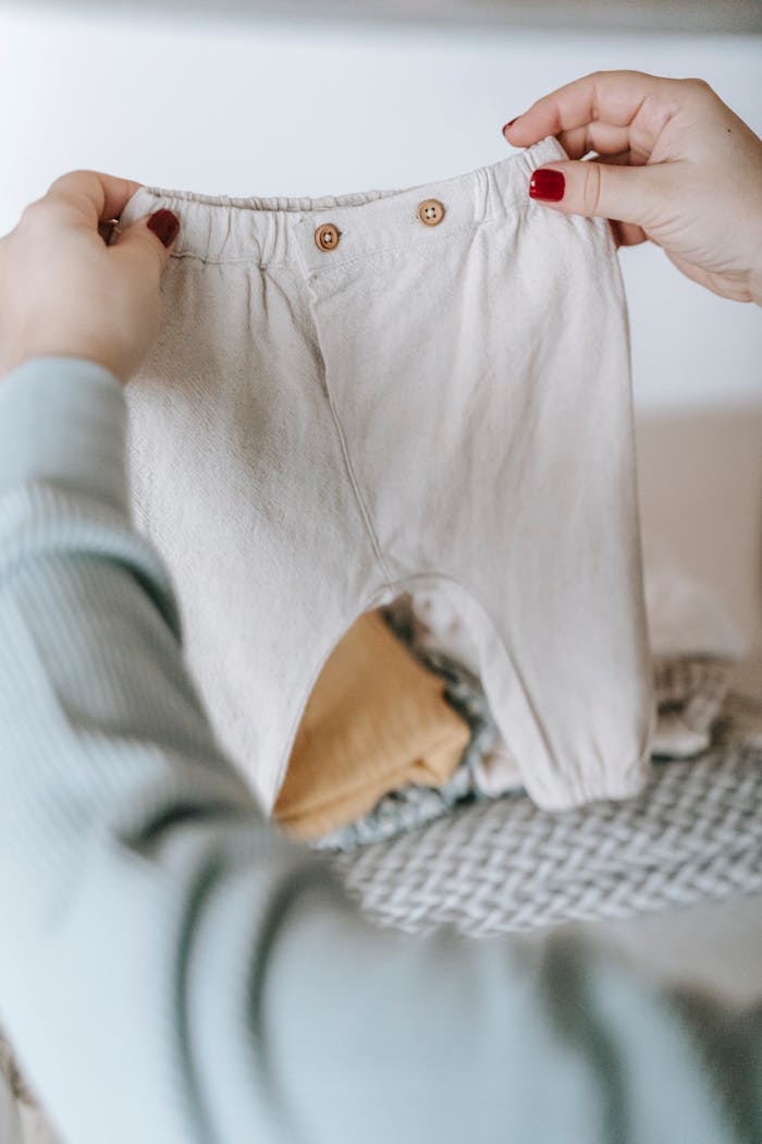 Crop faceless female with manicure and baby clothes in hands sitting at white table with garments in light room at home